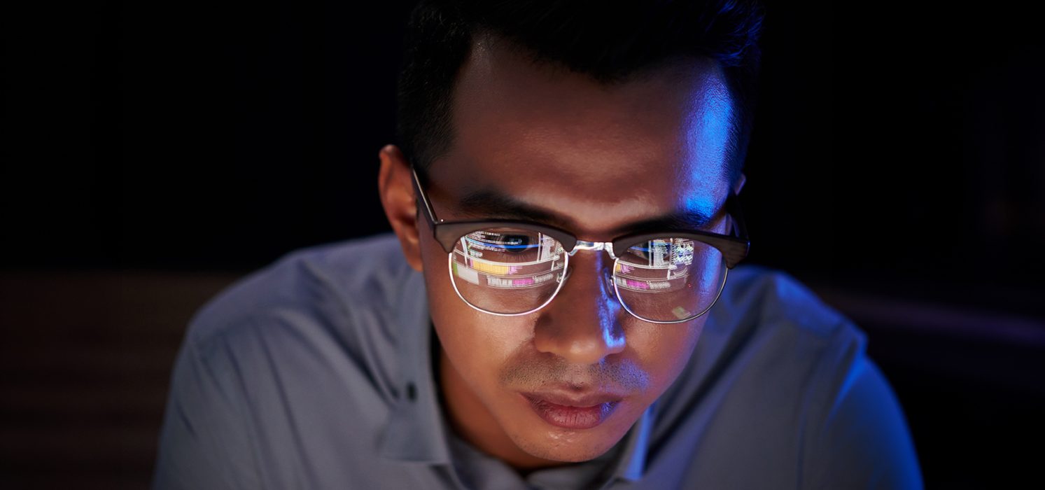 A man wearing glasses focused on a screen with reflections of code visible in the glasses.
