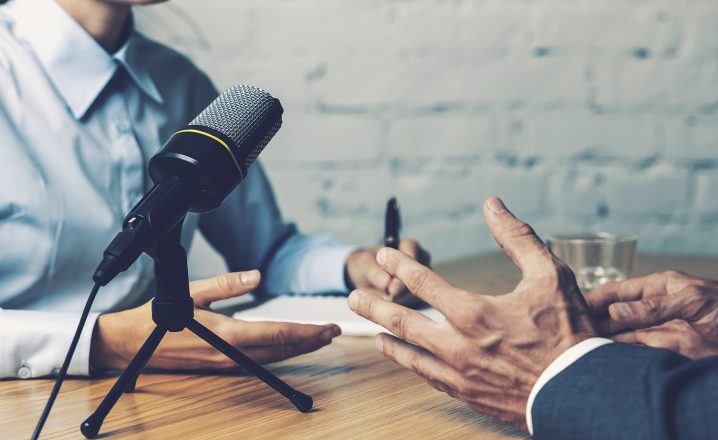 Pictorial representation of an interview between two individuals containing a microphone, their hands, a wooden table, and a white brick wall.