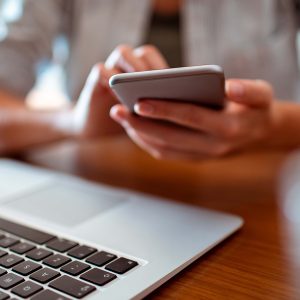 Pictorial representation of a laptop placed on a wooden table with a person's hand holding a mobile phone in the background.
