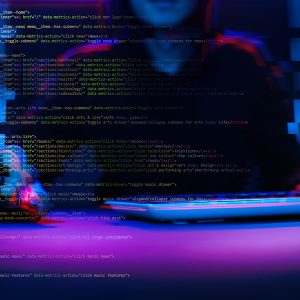Pictorial representation of phishing campaign. A blurred image focusing on a person typing on a laptop with lines of code visible on the screen, illuminated in blue and red lights, suggestive of intense coding or cyber activities.