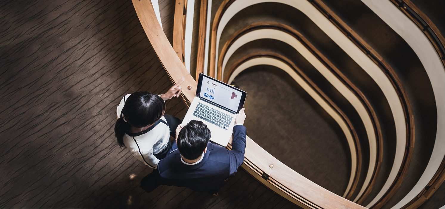 Two individuals are standing on a curved balcony inside a modern building, looking at a laptop displaying charts. The interior design features multiple layers with wooden railings and a central spiral staircase.