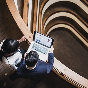 Two individuals are standing on a curved balcony inside a modern building, looking at a laptop displaying charts. The interior design features multiple layers with wooden railings and a central spiral staircase.