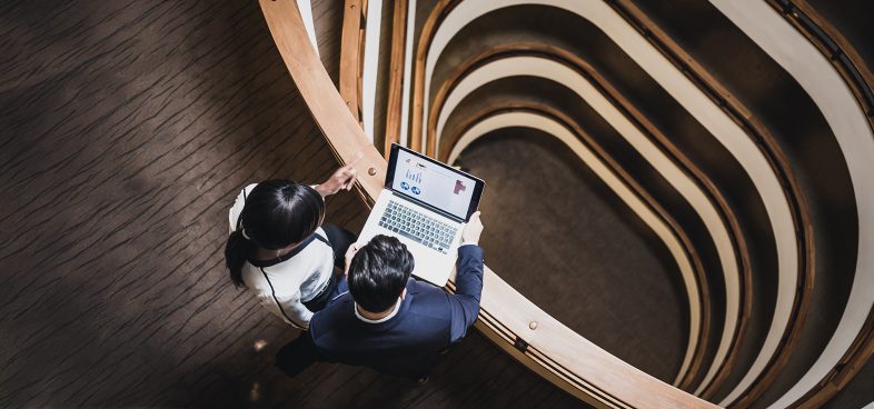 Two individuals are standing on a curved balcony inside a modern building, looking at a laptop displaying charts. The interior design features multiple layers with wooden railings and a central spiral staircase.