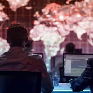 Pictorial representation of security risks in AI models. A group of people is seated at computer workstations in a dimly lit room, facing a large screen displaying a world map with data visualizations.