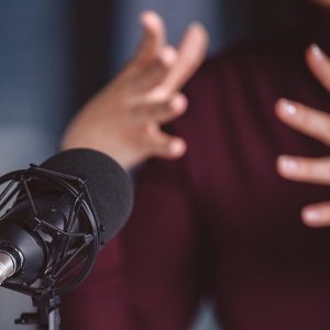 Pictorial representation of a microphone with a blurred background of an individual wearing a maroon shirt moving their hands expressively.