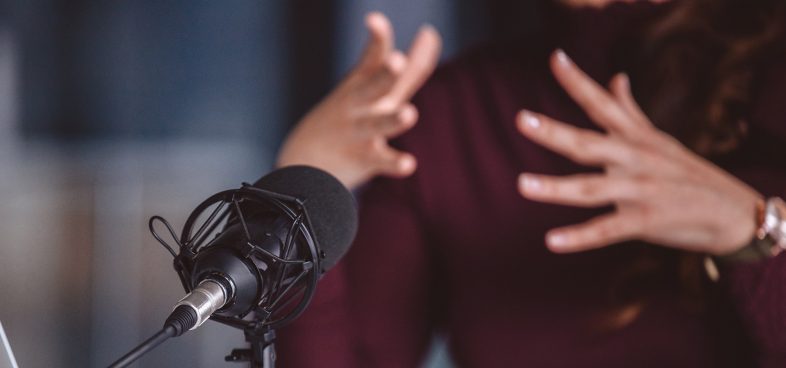 Pictorial representation of a microphone with a blurred background of an individual wearing a maroon shirt moving their hands expressively.