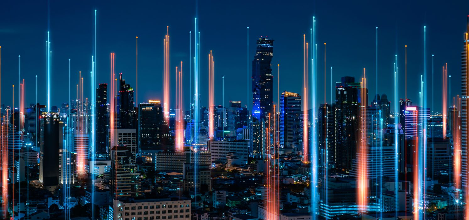 Pictorial representation of large-scale credential attacks. A vibrant city skyline at night, with tall skyscrapers and glowing digital beams extending into the sky, suggesting advanced technology and connectivity.
