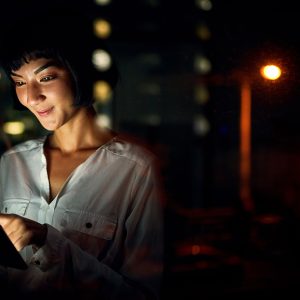 Pictorial representation of a female smiling while looking at the screen of a tablet. The backgorund is dark and features warm-toned lights.